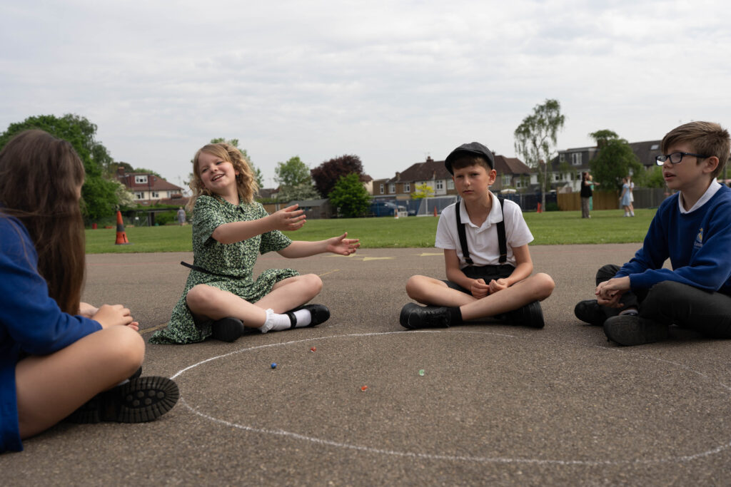Children dressed in period costumes (possibly 1930s or wartime era) are playing marbles outdoors. This suggests a re-enactment or an activity designed to reflect the school's past.