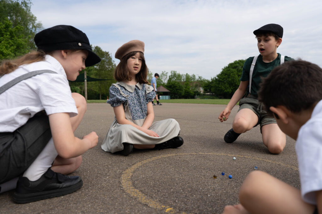 Children dressed in period costumes (possibly 1930s or wartime era) are playing marbles outdoors. This suggests a re-enactment or an activity designed to reflect the school's past.