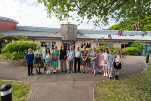 A group photo outside a building, likely the school. It includes both adults and children, with some children dressed in what appear to be historical costumes (e.g., flat caps, suspenders) and Union Jack bunting visible in the background, further supporting a celebratory, historical theme.