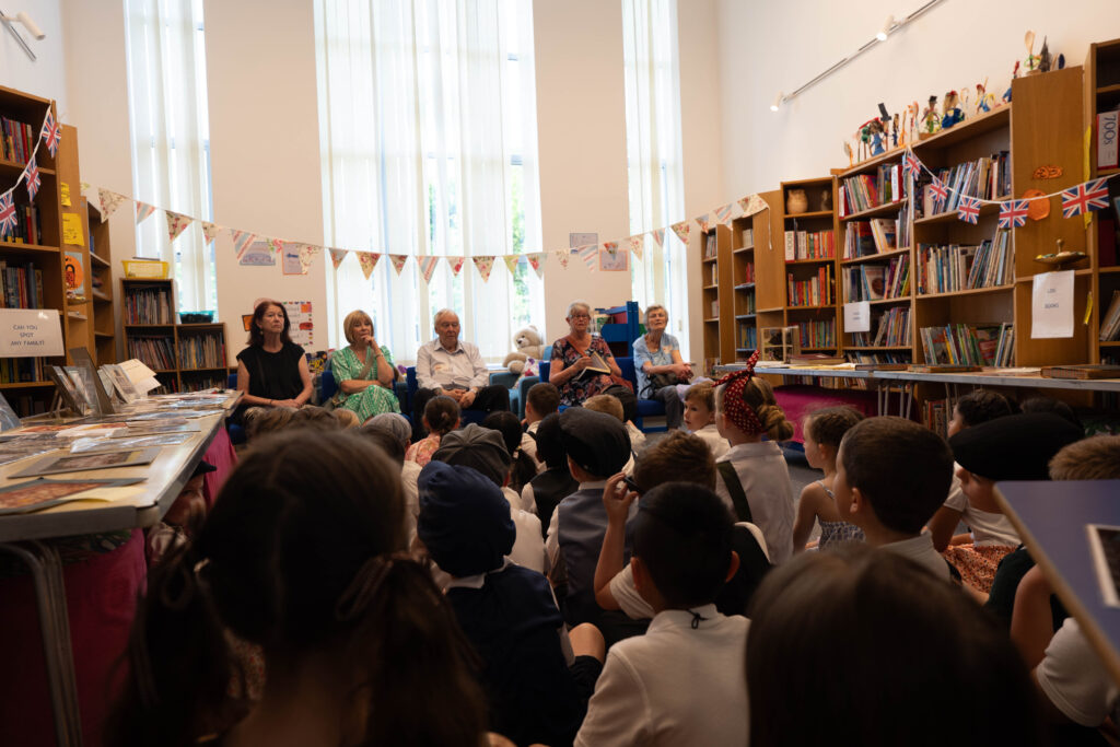 Children are seated on the floor in a library or classroom setting, attentively listening to adults.