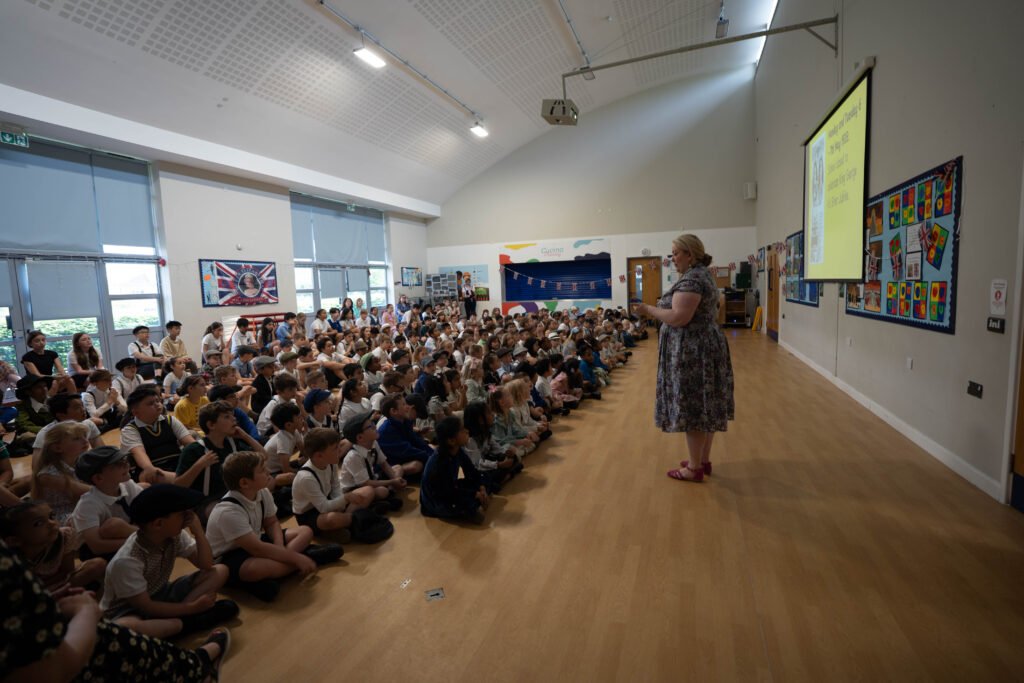 A large group of children, many in historical dress, are seated on the floor in what looks like a school hall, listening to an adult woman who is standing and presenting. This could be a historical talk or assembly related to the school's history.
