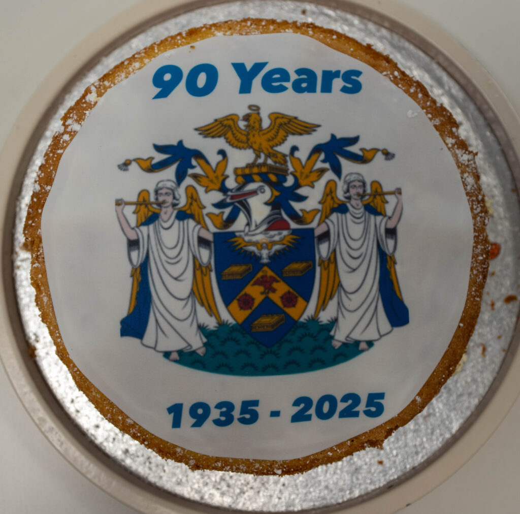 This image shows a cake celebrating "90 Years" and the dates "1935-2025," strongly indicating an anniversary event. The emblem on the cake might be the school's crest or a local coat of arms.
