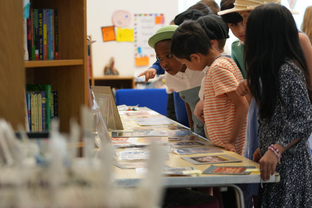 Children are looking at historical documents or photographs displayed on a table. This is likely an exhibition or display designed to educate them about the school's history.