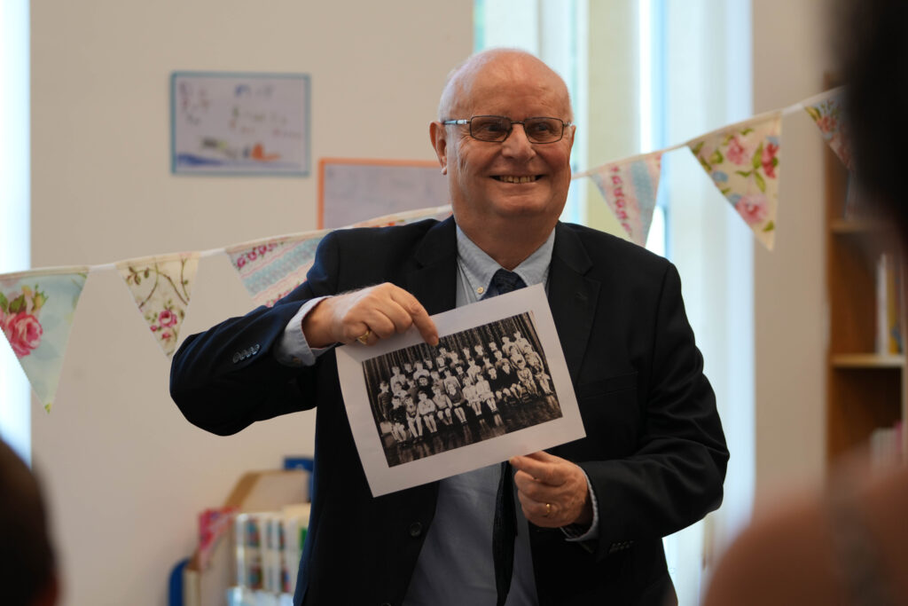 An older gentleman is holding up a black and white photograph of a group of children, presumably an old school photo. He's smiling, indicating a positive memory or connection to the past. This reinforces the historical theme.
