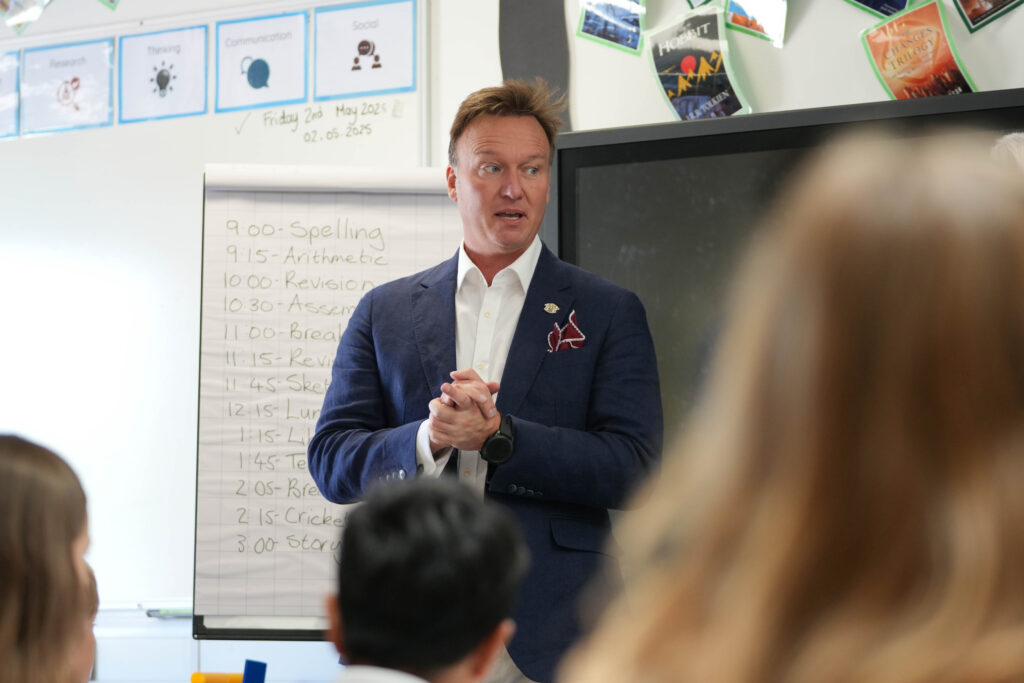 An adult man is speaking to a group of children in a classroom. A whiteboard behind him shows a schedule for "Friday 2nd May 2025" with activities like "Spelling," "Arithmetic," "Revision," "Assembly," and "Cricket." While this specific image doesn't directly show historical elements, it places the event within a school context, likely part of the overall anniversary celebrations.