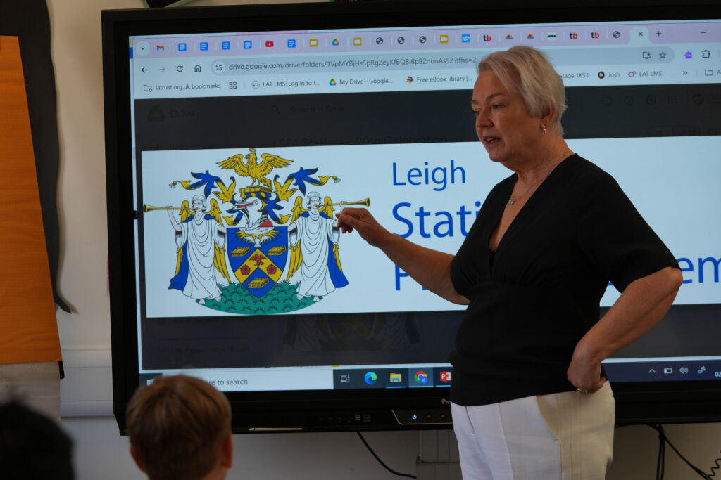 This image shows an adult woman, likely a teacher or school official, pointing to a large screen displaying a coat of arms or emblem. The text visible on the screen partially reads "Leigh Stationers' Primary," suggesting this is the school's official emblem.
