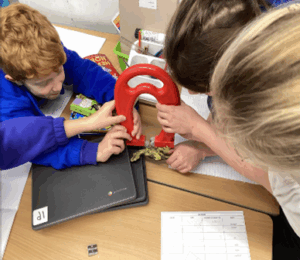A close-up of students using a large red horseshoe magnet to experiment with small metallic objects on a desk.