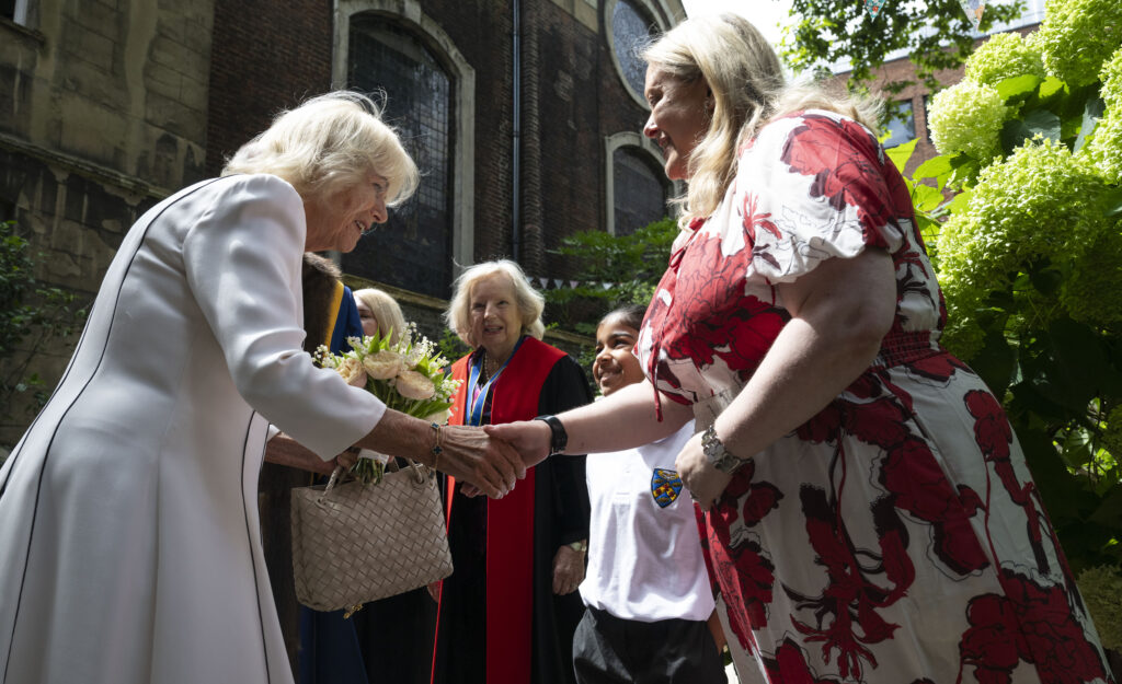 Image 1 (ly32_15-7-25_Queen_Stationers.jpg): This image captures a moment during Queen Camilla's installation as an Honorary Freeman and Liveryman of the Worshipful Company of Stationers and Newspaper Makers on July 15, 2025. Queen Camilla, on the left, is seen shaking hands with a woman in a red and white floral dress. Queen Camilla is holding a small bouquet of light-colored flowers. In the background, to the right of Queen Camilla, an older woman in academic-style robes (red with blue trim) is visible, along with a younger person in a school uniform, both looking towards the interaction. The setting appears to be outdoors, with a building made of brick in the background to the left and green foliage on the right.