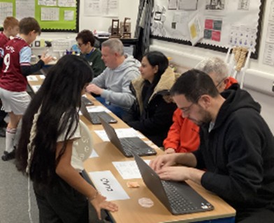 A candid photograph of a busy classroom or community center setting. Several adults and children are seated or standing around a long table equipped with laptops, appearing to be engaged in a workshop or collaborative educational activity.